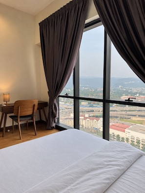 Ein Hotelzimmer mit einem großen Bett, einem Schreibtisch und einem Stuhl. Durch ein großes Fenster ist ein Blick auf die Stadt möglich.