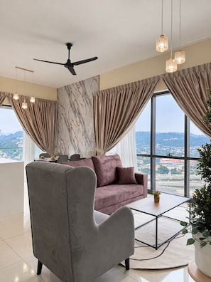 Ein modernes Wohnzimmer mit einem grauen Sessel, einem rosafarbenen Sofa und einem Couchtisch. Es gibt einen Deckenventilator, große Fenster mit Vorhängen und einen Blick auf eine Stadtlandschaft.