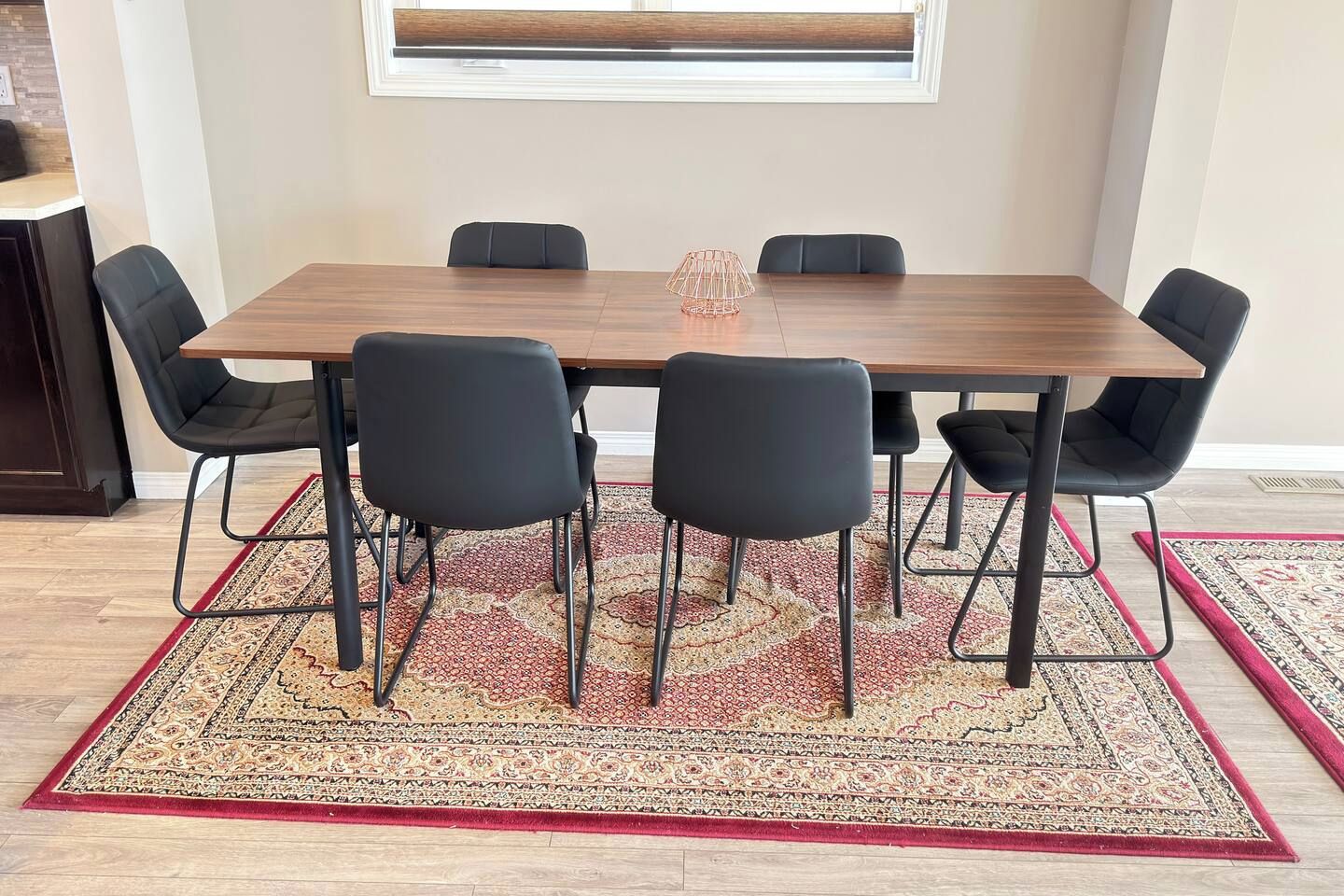 A dining area with a wooden table, six black chairs, a patterned rug, and a window above.