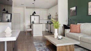 A modern kitchen with white cabinets, a central island, and a dining area with a white table and bench. - Landing | Stylish 1BD, Game Room, Gym (Gainesville)