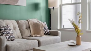 A living room with a sofa, pillows, a vase with flowers, a lamp, and a window with a view. - Landing | Modern 1BD, Gym, Pool (Gainesville)