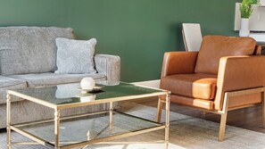 Ein modernes Wohnzimmer mit einem Glastisch, einer grauen Couch mit gemusterten Kissen und einem braunen Ledersessel.