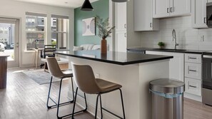 A modern kitchen with a central island, bar stools, a sink, and white cabinetry. - Landing | Stylish 1BD, Pool, Game Room (Gainesville)