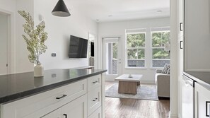 A modern kitchen with a black countertop, white cabinetry, a flat-screen TV, and a dining area with a wooden table and a sofa. - Landing | Chic 1BD, Game Room, Pool (Gainesville)