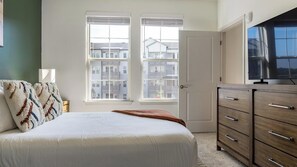 Ein Schlafzimmer mit einem Bett, einem Schminktisch, einem Fernseher und einem Fenster mit Blick auf Gebäude.