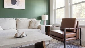 A room with a green wall, a white sofa, a brown leather chair, a white table, and a window with a view of trees. - Landing | Beautiful 2BD, Game Room, Pool (Gainesville)