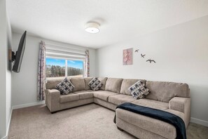 Un salon avec un canapé d’angle beige, un téléviseur à écran plat fixé au mur, une fenêtre avec des rideaux et un luminaire au plafond.