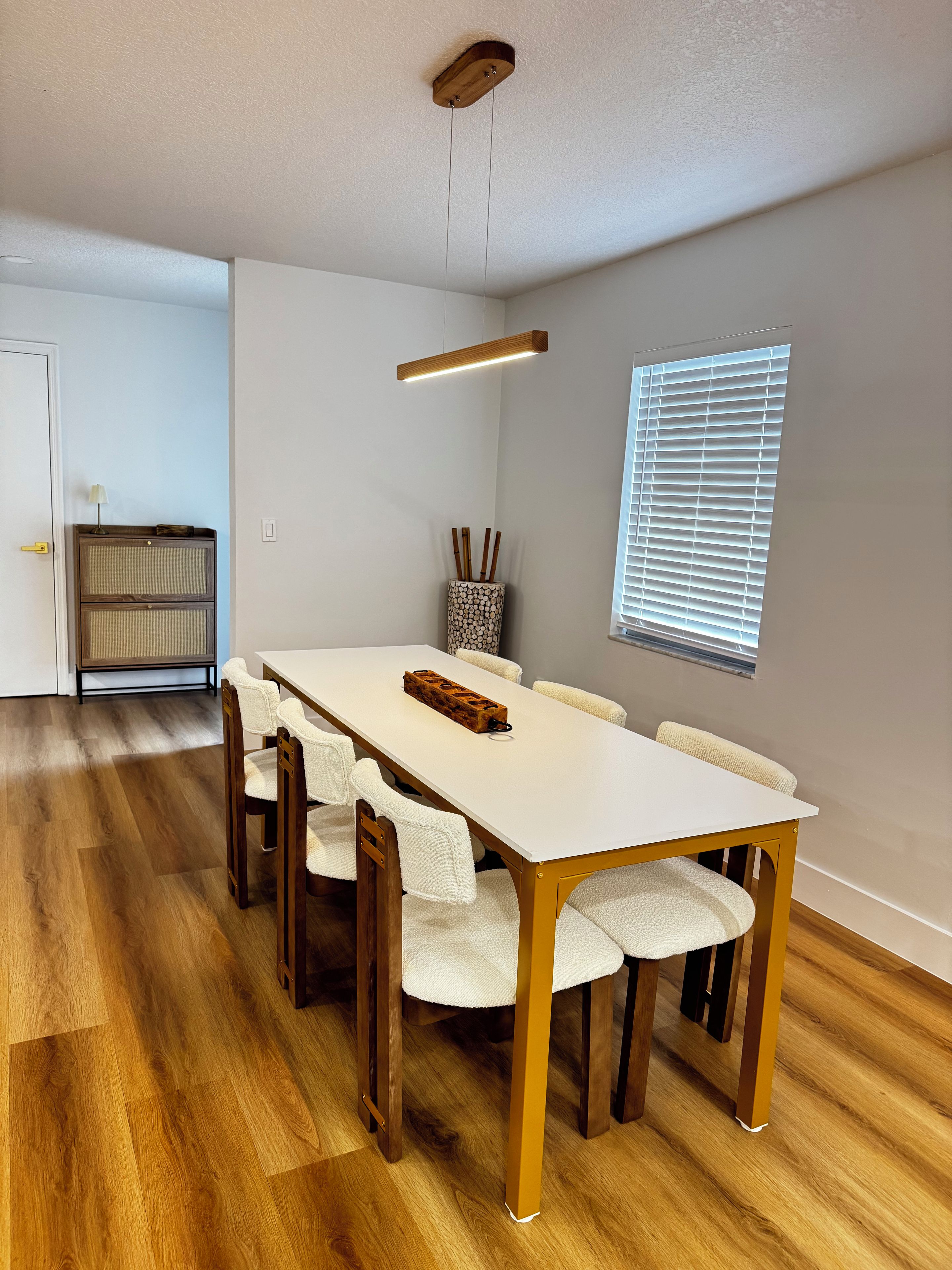 Een eetgedeelte met een houten tafel en stoelen, een plafondlamp en een raam met zonwering.