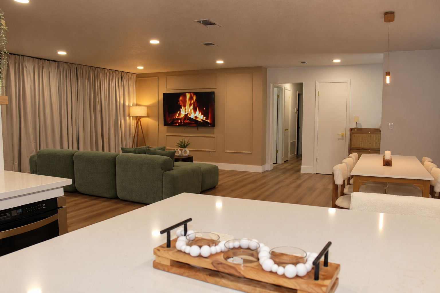A modern living room with a flat-screen TV, green sofas, a dining table with chairs, and a kitchen area with a dishwasher.