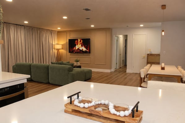 A modern living room with a flat-screen TV, green sofas, a dining table with chairs, and a kitchen area with a dishwasher.