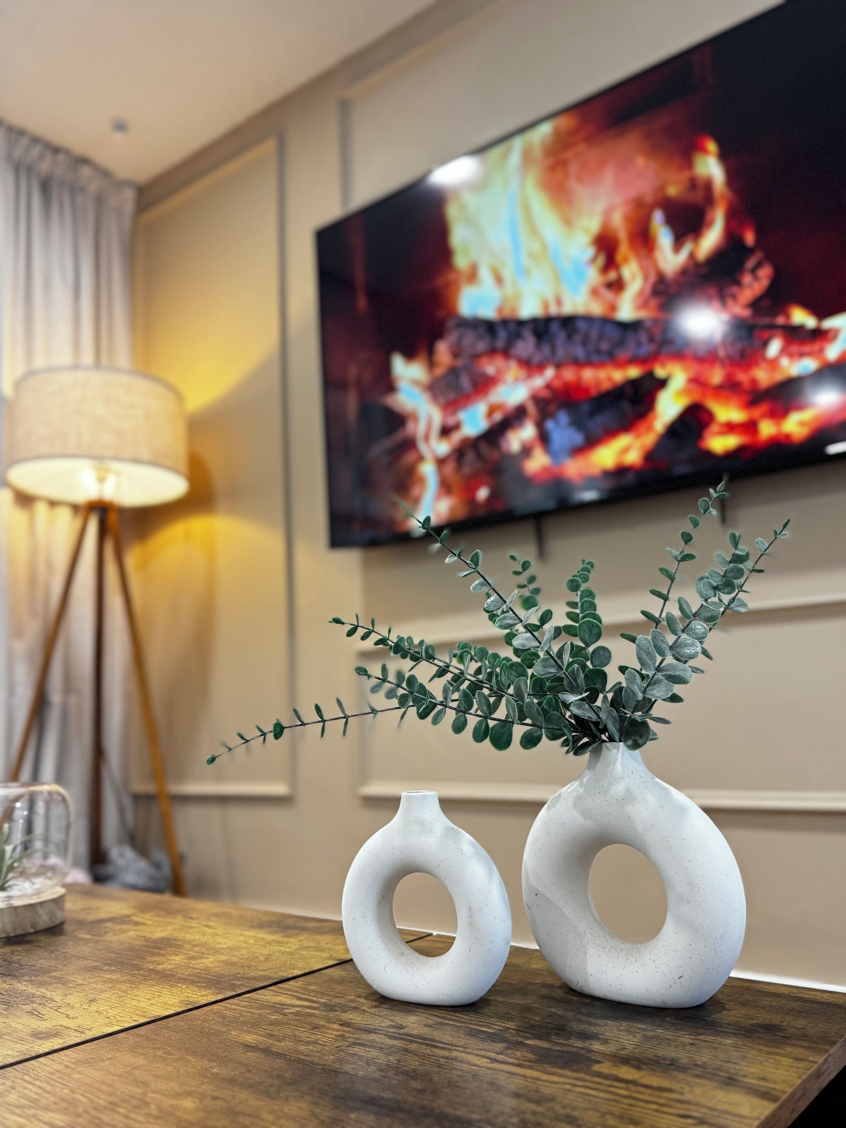 Een kamer met een houten tafel, twee witte vazen met ronde openingen, een vloerlamp en een televisie waarop een haard zichtbaar is.