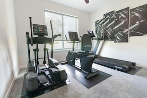 Un gimnasio en casa con cinta de correr, elíptica y bicicleta estática, un ventanal grande y una pared decorada con arte abstracto.