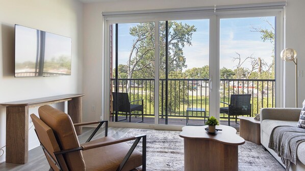 A modern living room with a large sliding glass door leading to a balcony, a flat-screen TV mounted on the wall, and a wooden coffee table.