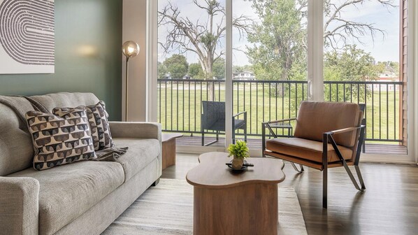 A living room with a sofa, a chair, a coffee table, and a view of a balcony with a railing and trees.