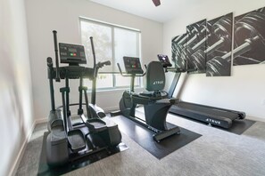 Een sportschool thuis met een loopband, een elliptische trainer en een hometrainer, een groot raam en een muur met abstracte kunst.