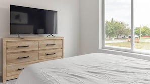 A bedroom with a bed, a wooden dresser, a television on top, and a window with a view of the outside. - Landing | Incredible 1BD, Gym (Council Bluffs)