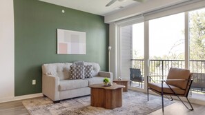 A living room with a sofa, coffee table, and chair, a painting on the wall, and a view through a sliding glass door. - Landing | Modern 2BD, Gym (Council Bluffs)
