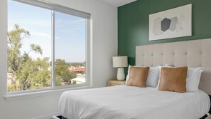 A hotel room with a large window, a bed with white bedding and two brown pillows, a nightstand with a lamp, and a green accent wall. - Landing | Modern 2BD, Gym (Council Bluffs)