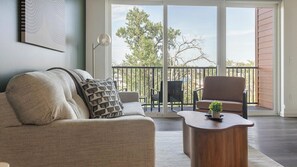 Een woonkamer met een bank, een stoel, een bijzettafeltje en uitzicht op een balkon met een hekwerk en bomen.
