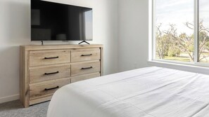Een slaapkamer met een bed, een houten dressoir met lades, een flatscreen televisie aan de muur en een raam met uitzicht op bomen.