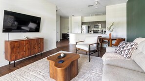 A modern living room with a flat-screen TV, wooden cabinet, white sofa, and a dining area with a table and chairs. - Landing | Stunning 1BD, Gym, Clubhouse (Jacksonville)