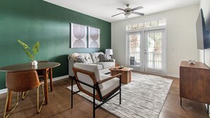 A living room with a green wall, a ceiling fan, a sofa, a coffee table, and a TV on a stand. - Landing | Stunning 1BD, Clubhouse, Gym (Jacksonville)