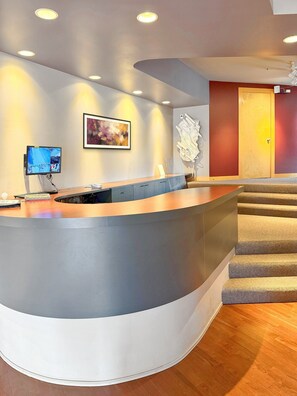 A reception area with a curved desk, a flat-screen monitor, a framed abstract painting, and a staircase leading to a door.