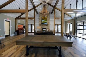A spacious indoor area with a high ceiling, wooden beams, a central chandelier, and a stone fireplace. - Landing | Modern 1BD, Gym (Bluffton)