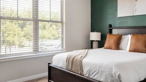A bedroom with a bed, a nightstand, a lamp, and a window with blinds. - Landing | Modern 1BD, Gym (Bluffton)