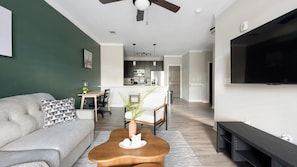 A modern living room with a sofa, coffee table, and TV. There is a kitchen in the background with a dining area. - Landing | Stunning 1BD, Gym (Bluffton)