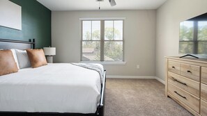 A bedroom with a bed, a nightstand, a lamp, a dresser, and a window with blinds. - Landing | Stunning 1BD, Gym (Bluffton)