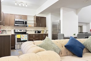 Una cocina moderna con electrodomésticos de acero inoxidable, una zona de comedor con una silla gris y un sofá beige con cojines.
