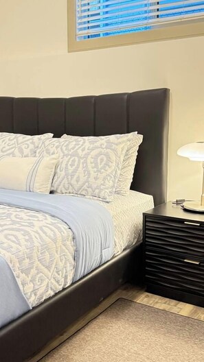 A bed with patterned bedding, a bedside table with a lamp, and a window with blinds.