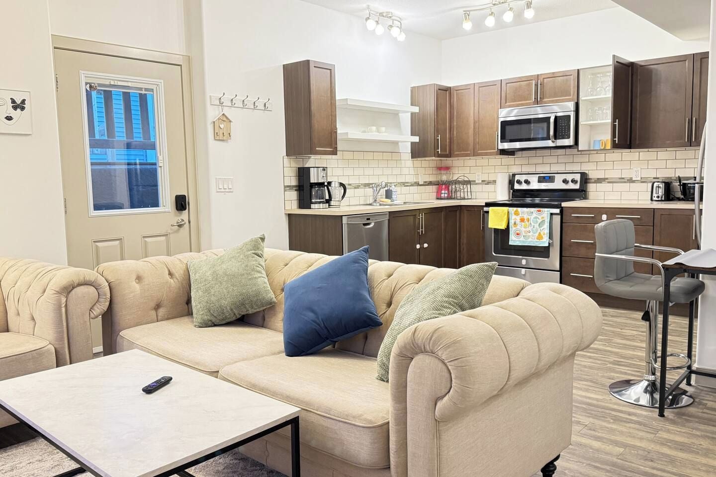 A compact living space with a sofa, a small table, a kitchenette with modern appliances, and a window with blinds.
