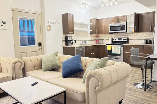 A compact living space with a sofa, a small table, a kitchenette with modern appliances, and a window with blinds.