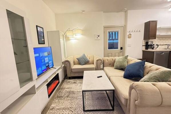 Una sala de estar moderna con un televisor de pantalla plana, un sofá color beige, una mesa de centro y una cocina con estufa y alacenas.