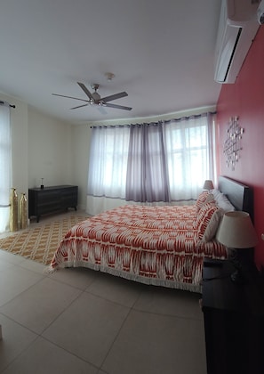 Ein Schlafzimmer mit einem Bett, einem Deckenventilator, einem Nachttisch, einer Lampe und einem Fenster mit Vorhängen.
