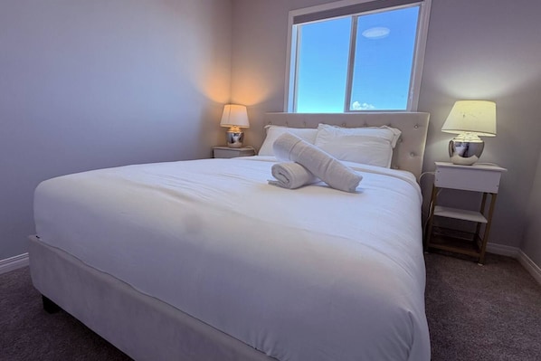 A neatly made bed with white linens, two bedside lamps, and a window with a view of the sky. - The Crossing Haven | 3BR Home (Calgary)