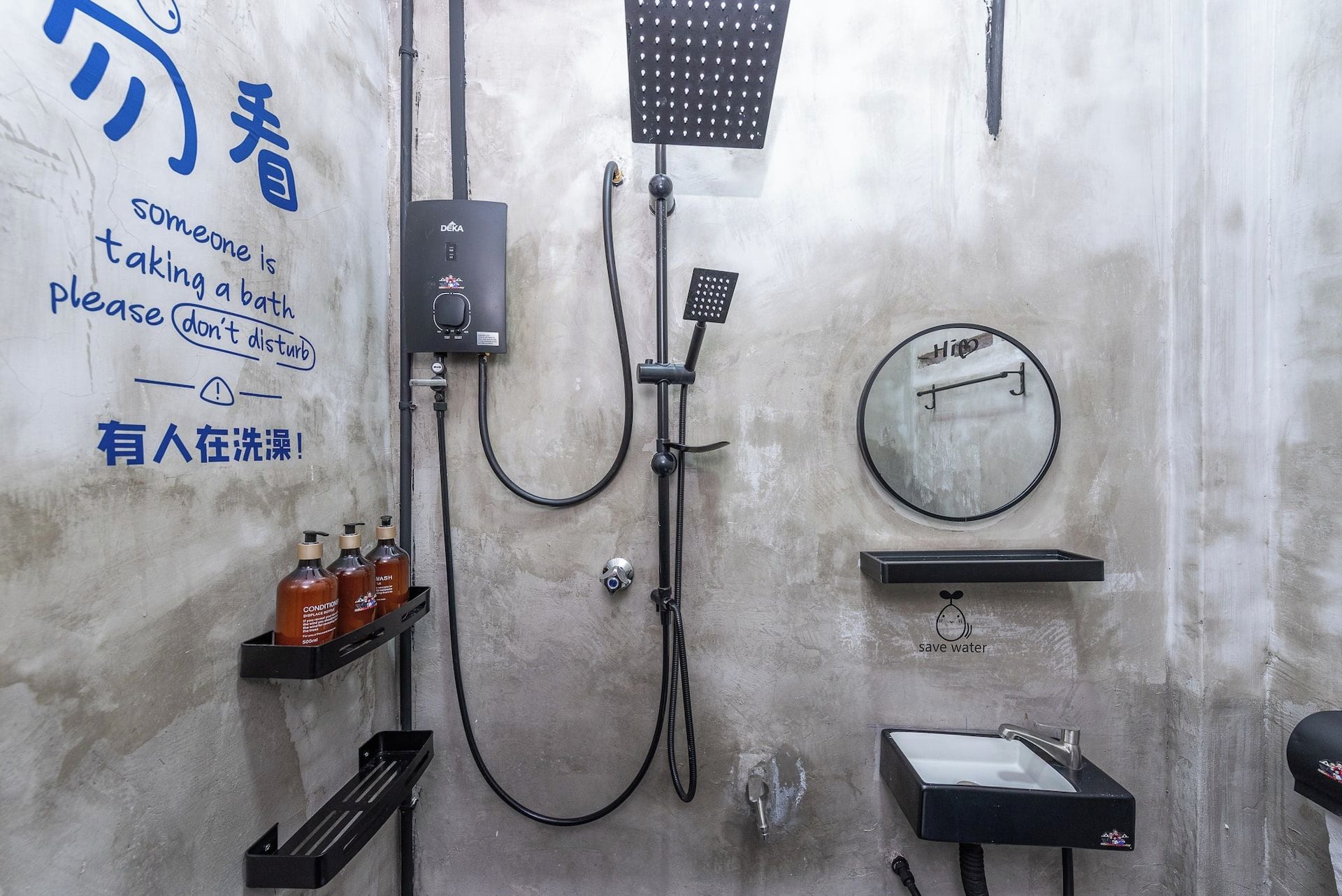 Un baño con ducha, lavamanos y espejo. Hay botellas de champú y acondicionador en una repisa. Un letrero en la pared solicita no molestar a alguien que se está bañando.