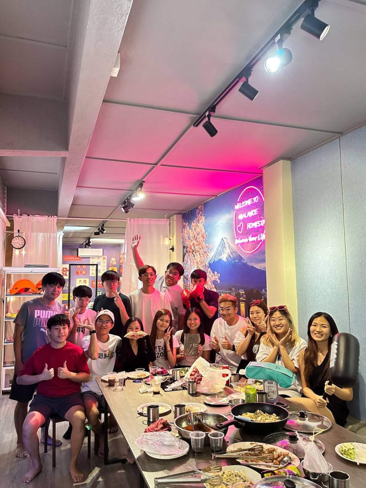 Un gruppo di persone sedute attorno a un tavolo con diversi piatti, in una stanza con un neon e un orologio appeso al muro.