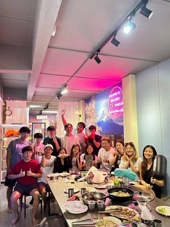 A group of people seated around a table with various dishes, in a room with a neon sign and a clock on the wall.