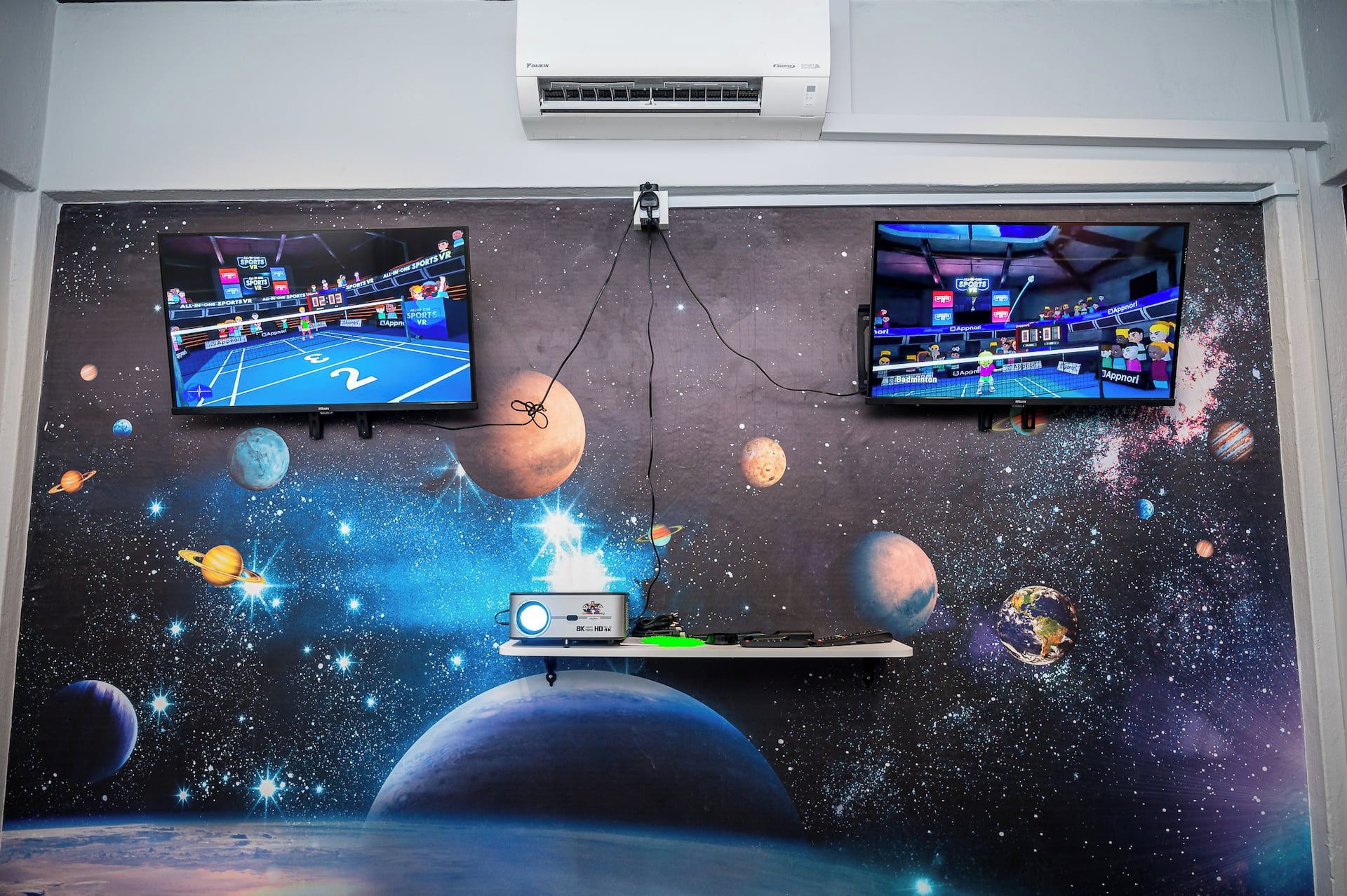 Two wall-mounted TVs displaying sports VR games, a projector, and a space-themed mural.