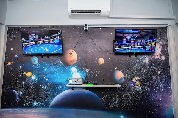 Two wall-mounted TVs displaying sports VR games, a projector, and a space-themed mural.