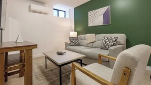 A living room with a green accent wall, a grey sofa, a wooden coffee table, and a small table with a remote control. - Landing | Chic 1BD, Gym (Bridgeport)