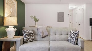 A living room with a grey sofa, patterned cushions, a white vase, and a lamp on a wooden table. - Landing | Chic 1BD, Gym (Bridgeport)