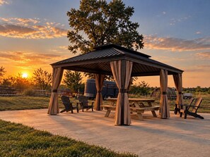 A gazebo with a table and chairs, surrounded by grass and trees, under a sunset sky.