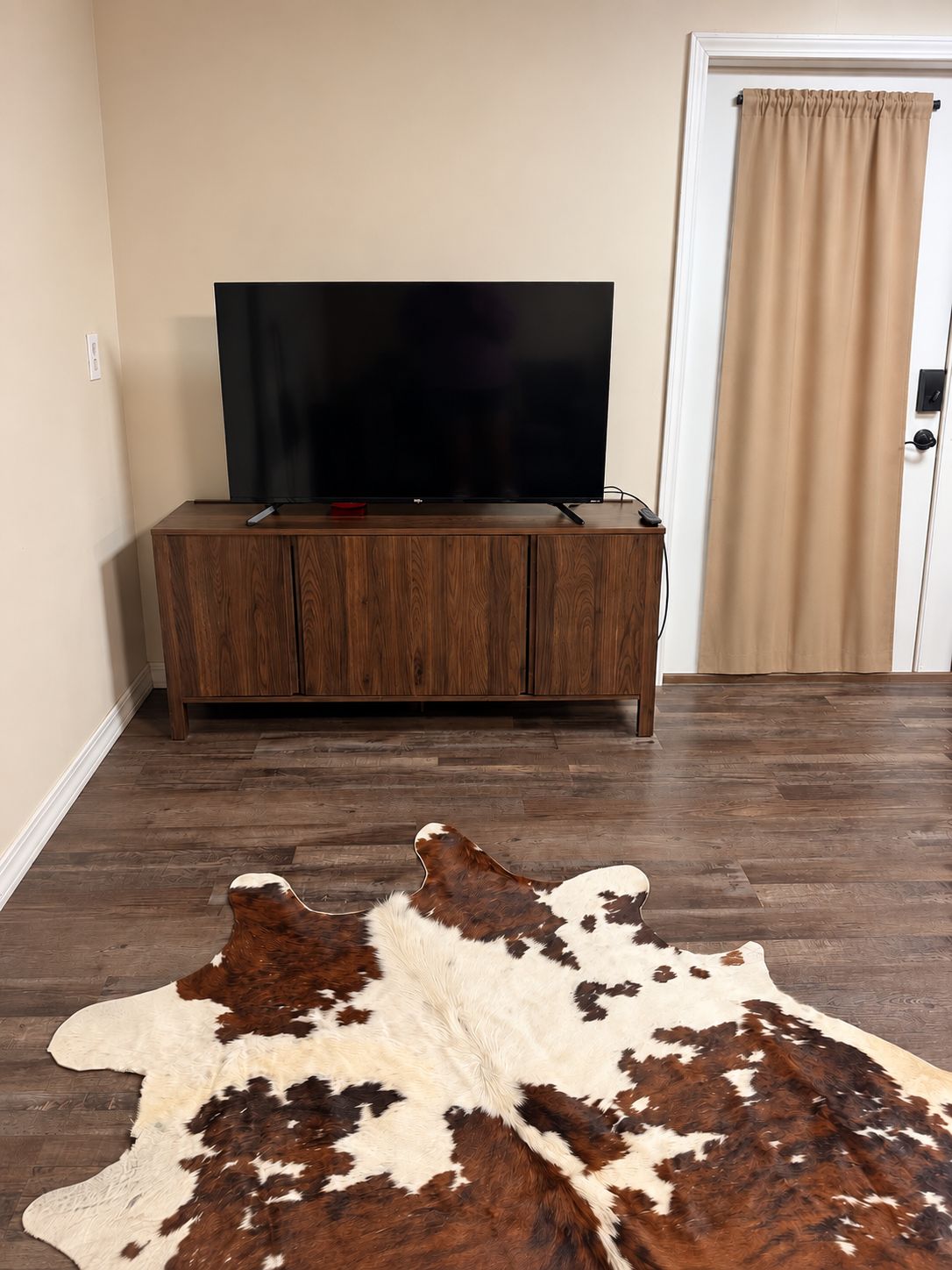 Une pièce avec un téléviseur à écran plat posé sur un meuble en bois, une porte avec des rideaux beiges et un grand tapis en peau de vache sur le sol.
