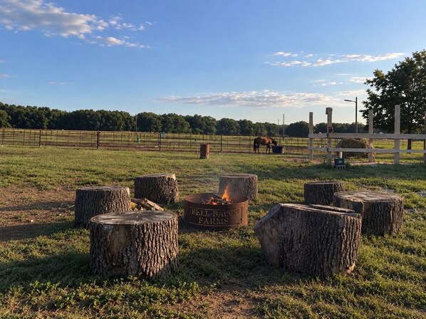 Ambiente rurale all'aperto con un braciere circondato da ceppi d'albero, un cavallo sullo sfondo e un'area recintata.