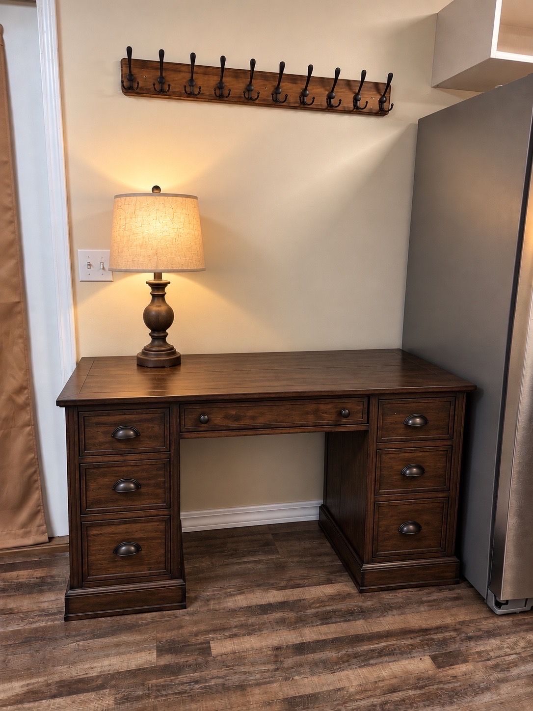 A wooden desk with drawers and a lamp, a coat rack on the wall, and a refrigerator in the corner.