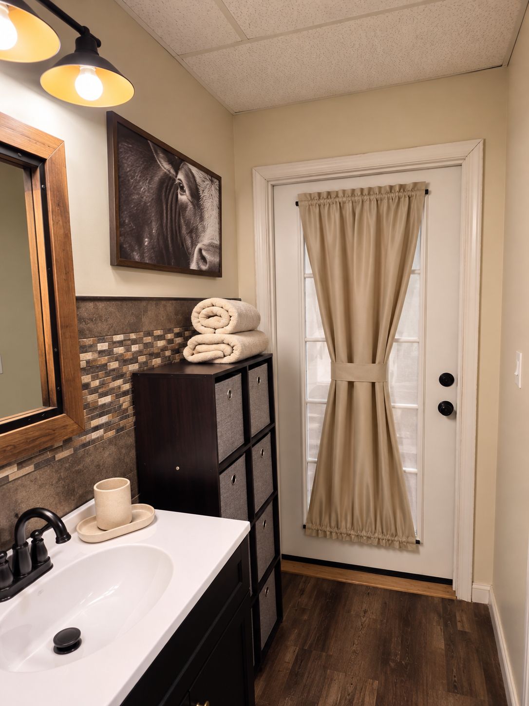 A bathroom with a white sink, dark wooden cabinets, a mirror, a towel rack, and a beige curtain.